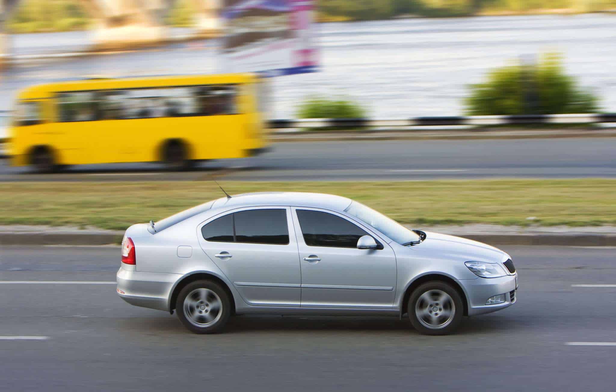 Silver car moving on a road with a yellow bus in the background.